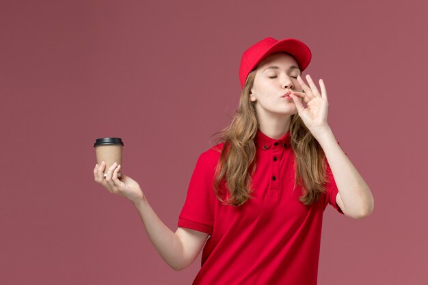 female courier in red uniform holding brown coffee cup showing tasty sign on light-pink, job uniform service worker delivery