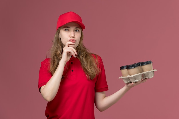 Free Photo female courier in red uniform holding coffee cups and thinking on pink, uniform job worker service delivery