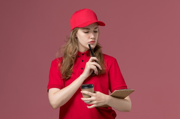 Free photo female courier in red uniform holding notepad pen and coffee cup on pink, uniform service delivery job worker