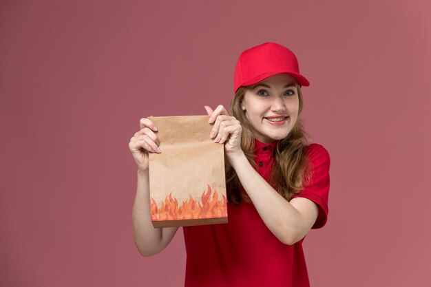 female courier in red uniform holding paper food package with smile on pink, job uniform worker service delivery