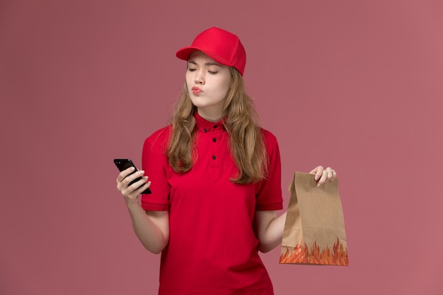 female courier in red uniform holding phone and food package on light-pink, job uniform service worker delivery