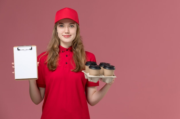 Free Photo female courier in red uniform smiling holding notepad and coffee cups on pink, uniform service delivery worker job