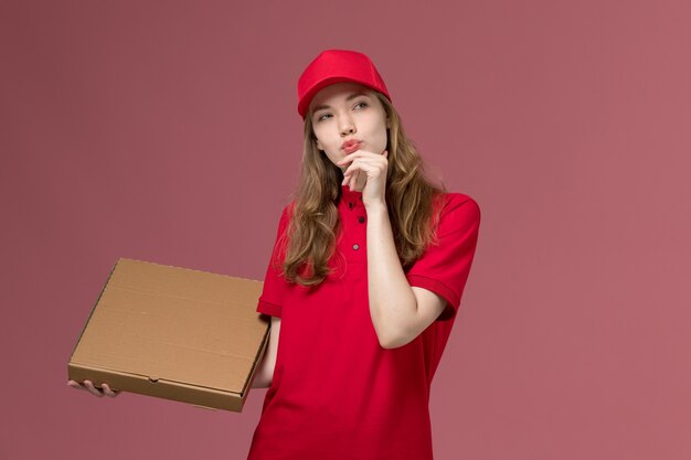 female courier in red uniform thinking holding food box on pink, uniform service delivery job