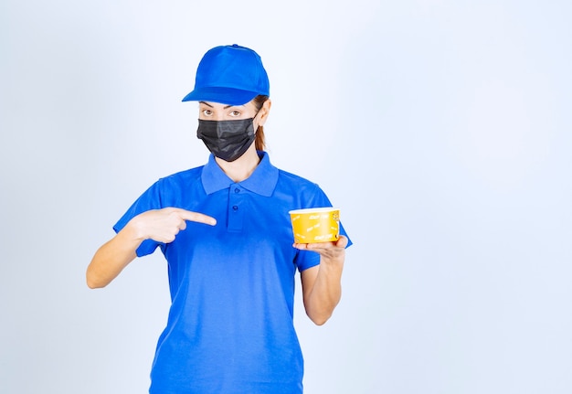 Female courier of the restaurant in blue uniform and face mask holding a takeaway food . 