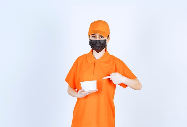 Free Photo female courier in yellow uniform and black mask holding a plastic cup and pointing at something.