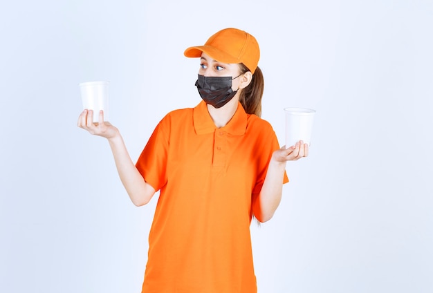 Female courier in yellow uniform and black mask holding takeaway drinks in plastic cups in both hands