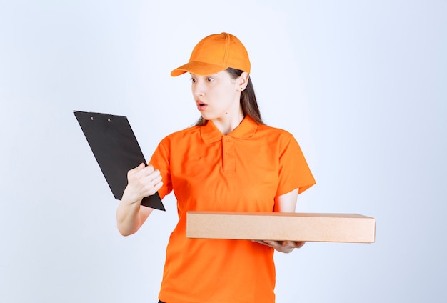 Female courier in yellow uniform holding a cardboard takeaway box and checking the address list and looks confused.