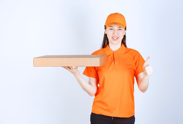 Female courier in yellow uniform holding a cardboard takeaway box and showing enjoyment hand sign.