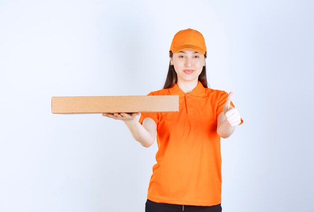 Female courier in yellow uniform holding a cardboard takeaway box and showing enjoyment hand sign.