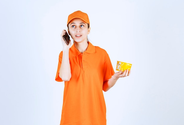 Female courier in yellow uniform holding a takeaway noodles cup and taking order from smartphone.