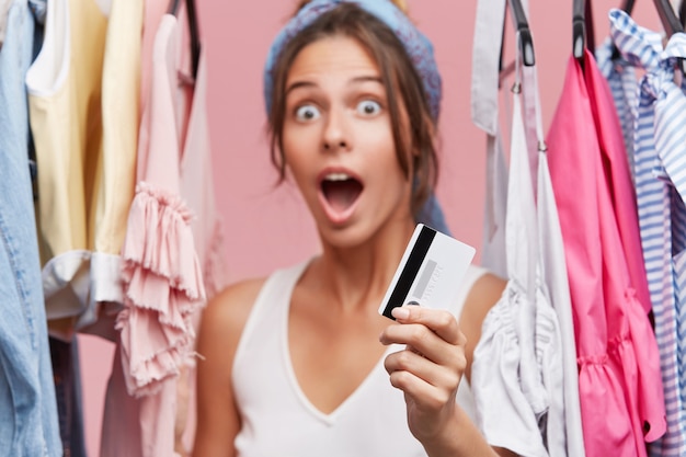 Female customer standing in boutique near rack with clothes, being surprised with high prices, holding plastic card in hand while having little money on her account, wanting to buy new dress