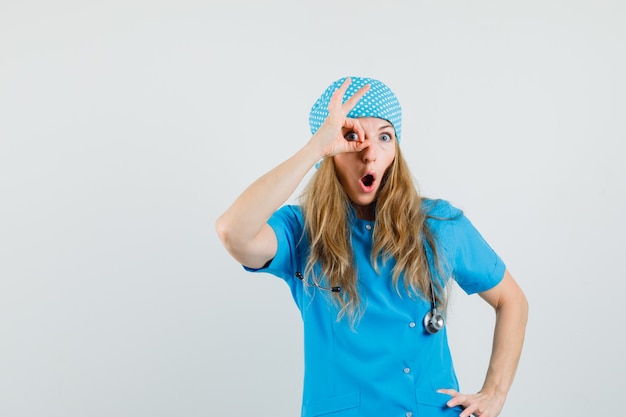 Free photo female doctor in blue uniform showing ok sign on eye and looking surprised