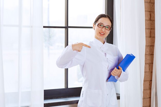 Female doctor in glasses holding blue folder near window. 