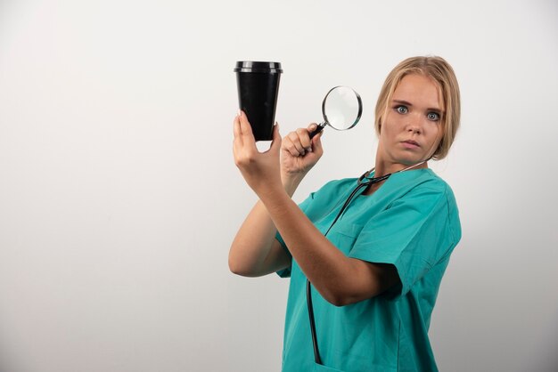 Female doctor holding cup and magnifying glass on white.