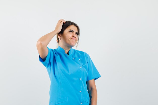 Female doctor scratching head while looking up in blue uniform and looking indecisive