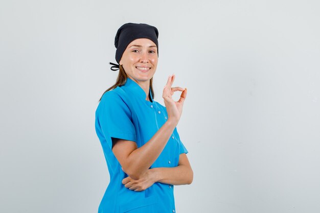 Female doctor showing ok sign in blue uniform, black hat and looking cheerful. front view.