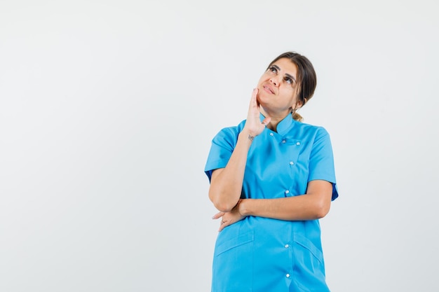 Female doctor standing in thinking pose in blue uniform and looking dreamy