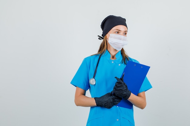 Female doctor in uniform, gloves, mask looking aside with clipboard and pen