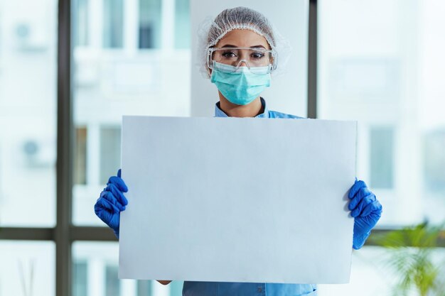 Female doctor with protective workwear holding empty cardboard while standing at clinic and looking at camera Copy space