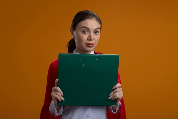 Free photo female doctor with stethoscope in white medical uniform and red superhero cape holding folder in front of her looking at front with serious face standing over orange wall