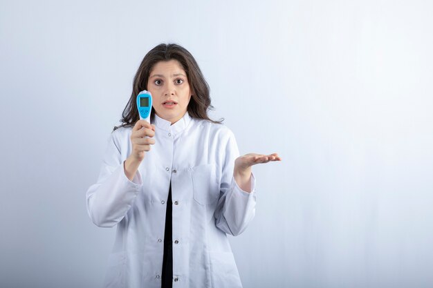 Female doctor with thermometer standing on white wall.