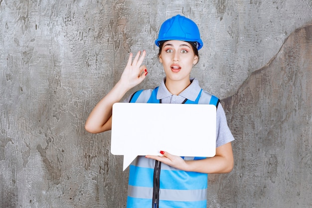 Free Photo female engineer in blue uniform and helmet holding a blank rectangle info board and showing enjoyment sign. 