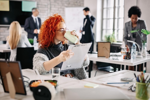 Free Photo female entrepreneur reading business reports and drinking coffee while sitting at her desk in the office there are people in the background