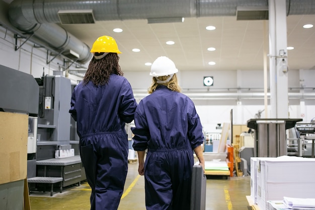 Female factory workers in hardhats and overalls walking on plant floor and talking, carrying toolkit box