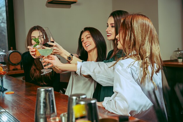 Female friends having a drinks at bar. They are sitting at a wooden table with cocktails. They are clinking glasses