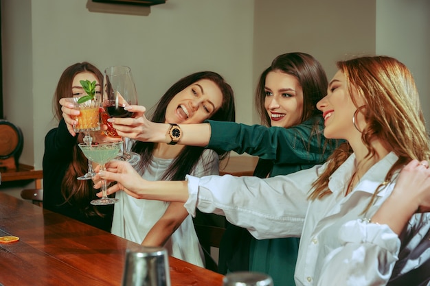 Female friends having a drinks at bar. They are sitting at a wooden table with cocktails. They are clinking glasses