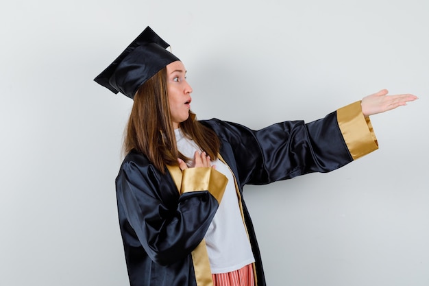 Free photo female graduate keeping hand on chest, stretching hand in academic dress and looking amazed. front view.