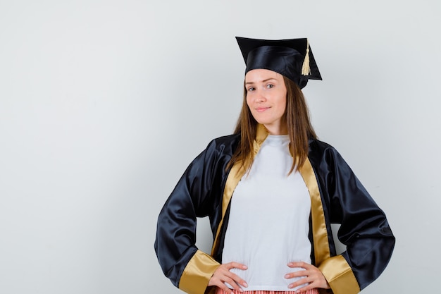 Free photo female graduate keeping hands on waist in academic dress and looking proud. front view.