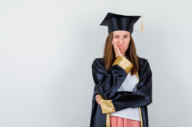 Female graduate leaning cheek on palm, curving lips in uniform, casual clothes and looking dissatisfied. front view.