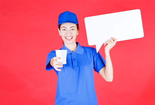 Female guide in blue uniform holding a white rectangular info board and offering a disposable cup of drink to the participant. 