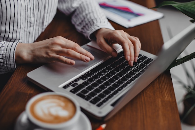 Female hands press laptop keys while sitting at wooden table with white cappuccino cup.