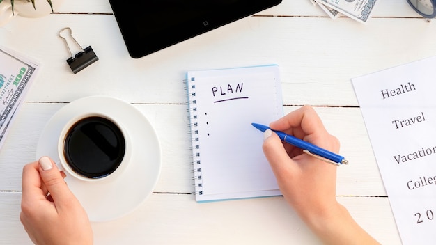 Female hands writing a to-do plan on a notepad, holding a coffee. Tablet, money. Wooden background