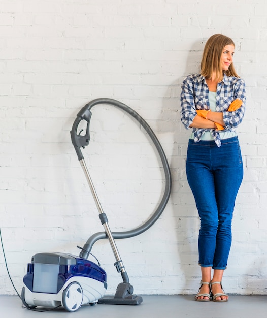 Free Photo female housemaid standing near vacuum cleaner in front of brick wall