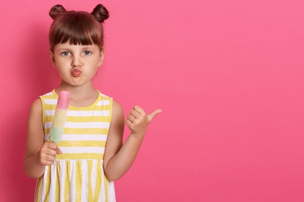 Female kid holding ice cream and pointing aside with thumb, posing isolated over rosy wall, keeping lips rounded, small girl looks businesslike and funny.