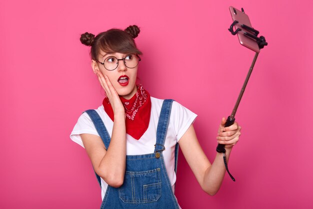 female making selfie, using selfie stick, poses, takes photo for her social network, makes surprised look, stands with open mouth, keeps hand on cheek. Teen concept.