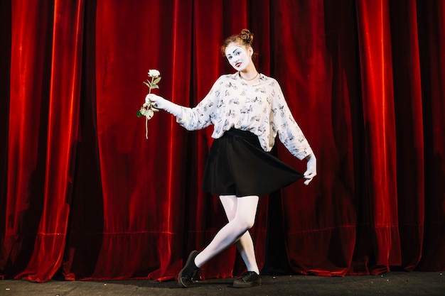 Free photo female mime artist standing with crossed leg holding white rose