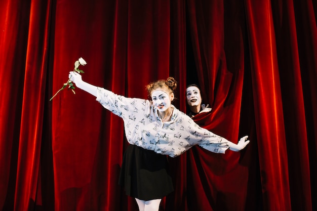 Free photo female mime holding white rose performing on stage with male mime peeking behind the curtain