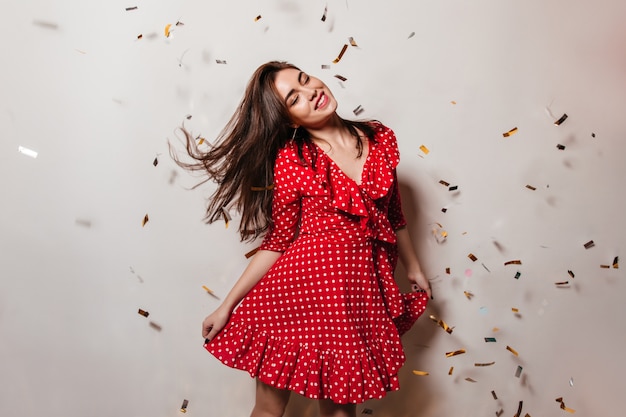 Female model closed her eyes from pleasure and dancing with smile on white wall with falling confetti. Photo of lady in red dress.