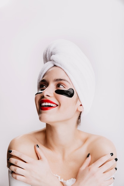 Female model with red lips looks upwards with enthusiasm. Girl in towel after shower posing on white wall.