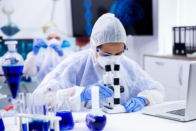 Female scientist looking through a microscope in research laboratory. Smoking blue liquid in test tubes.