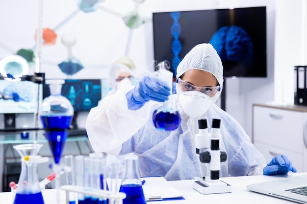 Female scientist in protection equipment holding and looking at a test tube with blue solution. Chemistry laboratory.