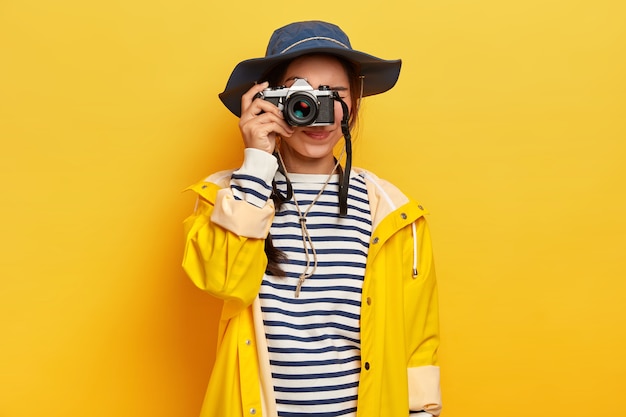 Female traveler makes memorable photos during trip, holds retro camera, takes images of beautiful landscape or place, dressed in striped jumper, raincoat and hat, isolated over yellow wall