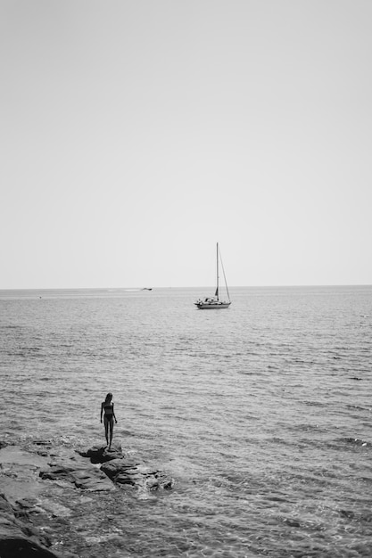 Free Photo female wearing bikini standing on a rock by the body of water with a sailboat floating in the sea