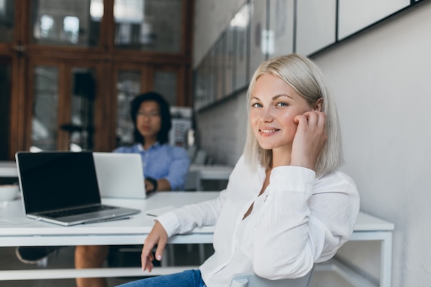 Female web-developer posing with smile in office while her asian male colleague working on project. Chinese marketer using laptop sitting at the table with pretty european manager.