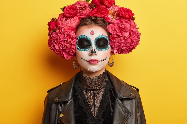 Free photo female with sugar skull drawn with smile, wears flower wreath, dressed in black clothes