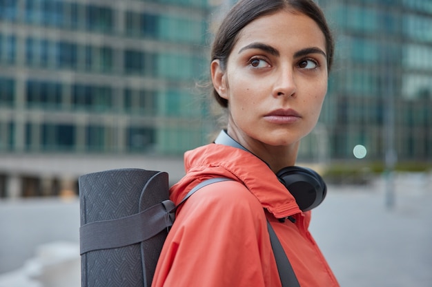 Female yoga trainer waits for someone at street has workout break restrores strength after training leads healthy lifestyle stands sideways against blurred city 